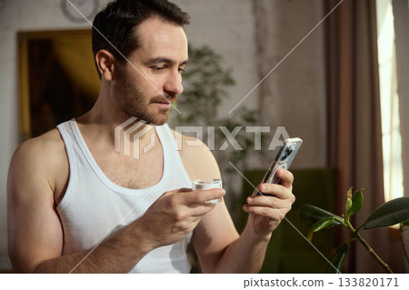 Man looking at phone while holding facial cream jar. Man looking at phone while holding facial cream jar. 133820171