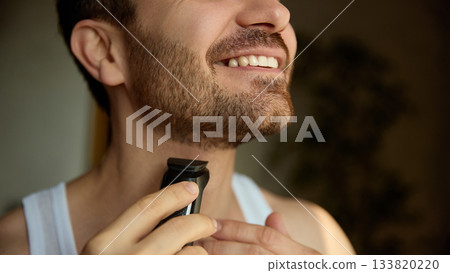 Smiling man trimming beard with electric grooming tool Smiling man trimming beard with electric grooming tool 133820220