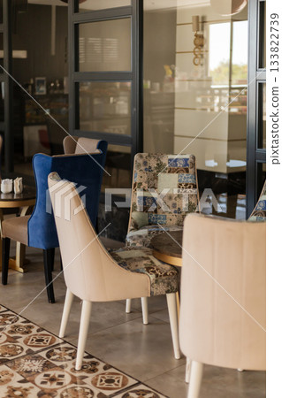 Cozy seating area features a mix of colorful and patterned chairs. Large windows offer natural light, enhancing the inviting atmosphere of the cafe 133822739
