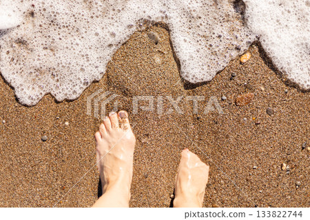 Warm sunlight shines on bare feet as they rest on soft sand, while gentle waves wash up and create a peaceful atmosphere at the beach. Perfect for relaxation and enjoyment Warm sunlight shines on bare feet as they rest on soft sand, while gentle waves wash up and create a peaceful atmosphere at the beach. Perfect for relaxation and enjoyment 133822744