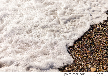 Gentle waves roll onto a sandy beach, creating a frothy foam along the shore. Small pebbles and grains of sand are visible as water recedes under the bright sun Gentle waves roll onto a sandy beach, creating a frothy foam along the shore. Small pebbles and grains of sand are visible as water recedes under the bright sun 133822745