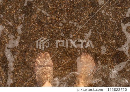 Bare feet stand in shallow water, feeling the gentle waves wash over them. Tiny pebbles and sand are visible under the clear water on a sunny afternoon Bare feet stand in shallow water, feeling the gentle waves wash over them. Tiny pebbles and sand are visible under the clear water on a sunny afternoon 133822750