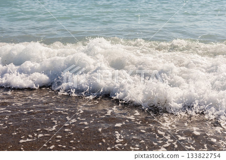 Gentle waves roll onto the shore, creating white foam as they reach the sandy beach. The sun reflects off the water, enhancing the serene summer atmosphere Gentle waves roll onto the shore, creating white foam as they reach the sandy beach. The sun reflects off the water, enhancing the serene summer atmosphere 133822754