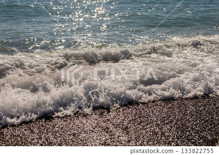 Waves roll onto a sandy beach under bright sunlight, creating a sparkling effect on the water. The tranquil scene invites relaxation and enjoyment of nature's beauty 133822755