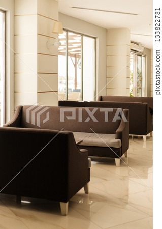 Cozy lounge area features several brown sofas arranged in an inviting manner. Natural light floods in through large windows, creating a warm atmosphere perfect for relaxation 133822781