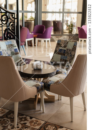A cozy seating area features two unique chairs and a round table. Bright natural light fills the space, creating a welcoming ambiance for visitors to relax or socialize A cozy seating area features two unique chairs and a round table. Bright natural light fills the space, creating a welcoming ambiance for visitors to relax or socialize 133822812