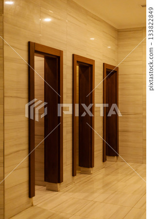 A serene hallway showcases three wooden door frames against a backdrop of smooth, light-colored marble. The warm lighting creates a calm atmosphere A serene hallway showcases three wooden door frames against a backdrop of smooth, light-colored marble. The warm lighting creates a calm atmosphere 133822849