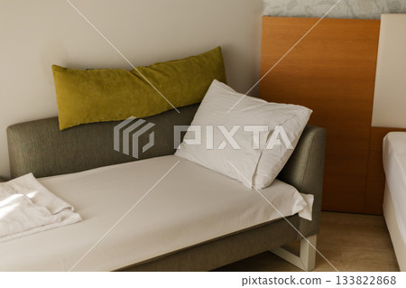 A cozy space features a minimalist bed with white linens and a grey sofa adorned with green throw pillows, creating a serene atmosphere perfect for relaxation A cozy space features a minimalist bed with white linens and a grey sofa adorned with green throw pillows, creating a serene atmosphere perfect for relaxation 133822868