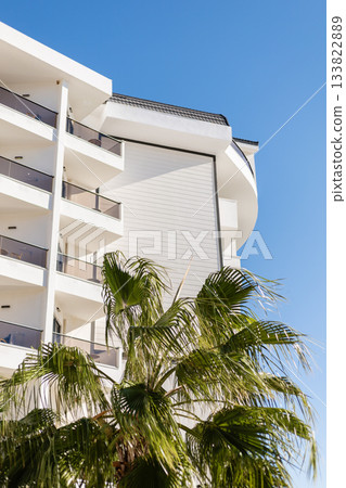 A contemporary building features multiple balconies and a palm tree in the foreground. The clear blue sky highlights the sunny atmosphere of this pleasant location 133822889