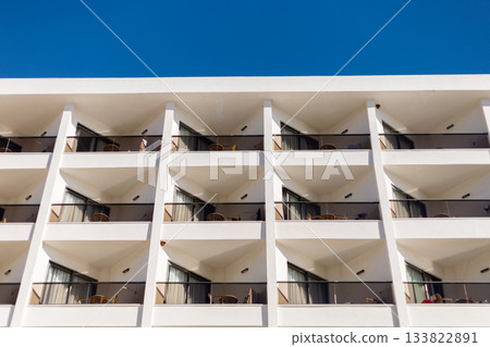 A modern hotel showcases its architectural design with multiple balconies facing a clear blue sky. Guests can enjoy the sunny weather and comfortable outdoor seating A modern hotel showcases its architectural design with multiple balconies facing a clear blue sky. Guests can enjoy the sunny weather and comfortable outdoor seating 133822891