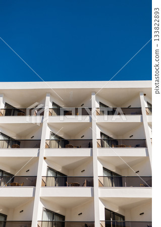 A contemporary hotel structure showcases multiple balconies with chairs, bathed in sunlight against a brilliant blue sky. The architecture highlights clean lines and open spaces A contemporary hotel structure showcases multiple balconies with chairs, bathed in sunlight against a brilliant blue sky. The architecture highlights clean lines and open spaces 133822893