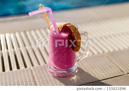 A vibrant pink drink is served in a clear glass, topped with a slice of tropical fruit and a straw. Sunlight shines down on the poolside, creating a perfect summertime vibe A vibrant pink drink is served in a clear glass, topped with a slice of tropical fruit and a straw. Sunlight shines down on the poolside, creating a perfect summertime vibe 133822896