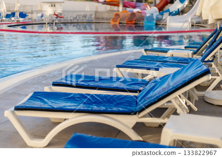 Blue loungers are arranged neatly beside a clean pool at a water park resort. Bright water slides are visible in the background, inviting guests to enjoy a day of fun and relaxation Blue loungers are arranged neatly beside a clean pool at a water park resort. Bright water slides are visible in the background, inviting guests to enjoy a day of fun and relaxation 133822967