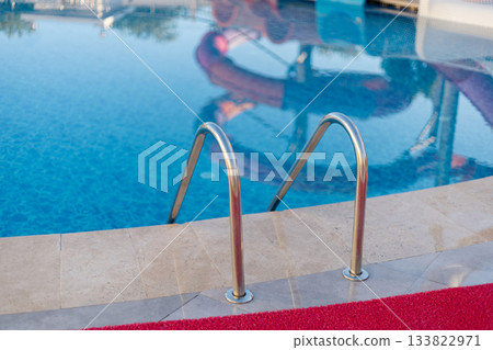 Bright blue waters reflect the sun, with shiny metal ladders positioned for easy access. Colorful swim floats add a playful touch to this inviting pool area 133822971