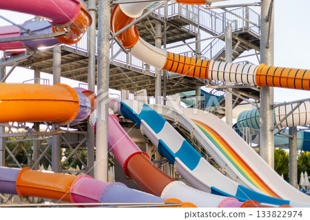A vibrant collection of water slides in various colors stands tall at a lively water park, inviting visitors on a warm summer day. Children and families are seen enjoying the fun 133822974