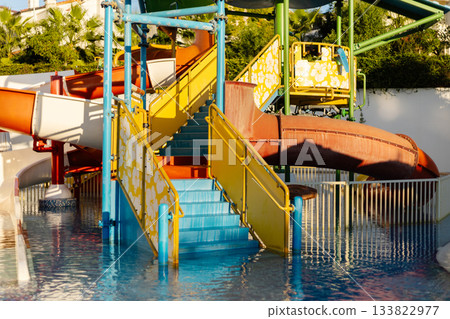Brightly colored water slides and a staircase invite visitors to enjoy fun and excitement at a water park. The atmosphere is lively under the warm sun and clear blue sky Brightly colored water slides and a staircase invite visitors to enjoy fun and excitement at a water park. The atmosphere is lively under the warm sun and clear blue sky 133822977