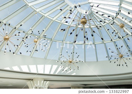 A bright interior showcases a modern ceiling design with a glass dome. Elegant starburst chandeliers hang from above, creating a stunning visual effect in the ample sunlight A bright interior showcases a modern ceiling design with a glass dome. Elegant starburst chandeliers hang from above, creating a stunning visual effect in the ample sunlight 133822997