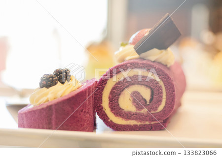 A beautifully arranged purple roll cake sits on a white plate, garnished with whipped cream, chocolate, and fresh blackberries. The cake showcases an inviting swirl design A beautifully arranged purple roll cake sits on a white plate, garnished with whipped cream, chocolate, and fresh blackberries. The cake showcases an inviting swirl design 133823066