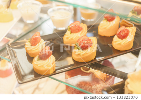 A selection of fresh, cream-filled strawberry desserts is beautifully arranged on a black tray in a bakery. The afternoon light accentuates the vibrant colors of the treats A selection of fresh, cream-filled strawberry desserts is beautifully arranged on a black tray in a bakery. The afternoon light accentuates the vibrant colors of the treats 133823070