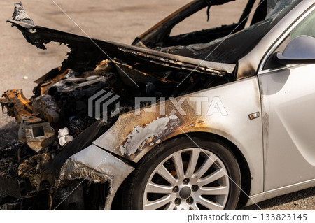 A silver car sits in an abandoned lot, heavily damaged by fire. Its burnt exterior reveals charred metal and melted parts. The sun shines down, highlighting the destruction A silver car sits in an abandoned lot, heavily damaged by fire. Its burnt exterior reveals charred metal and melted parts. The sun shines down, highlighting the destruction 133823145