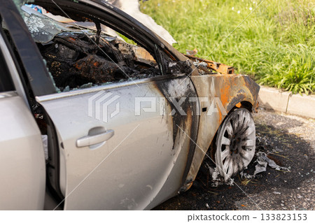 A car stands at the roadside, severely damaged by fire. Its body features burnt areas and a destroyed interior, with debris and ash around, reflecting a recent incident of a vehicle fire 133823153