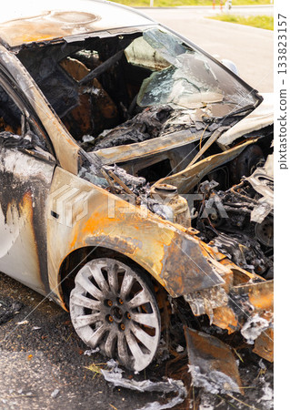 Burned car wreckage displays extensive damage with charred metal and ash. The vehicle rests by the road in an industrial area, showing signs of a recent fire 133823157