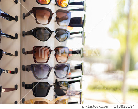 A selection of stylish sunglasses hangs on a display rack. The sun shines brightly, highlighting the different shapes and colors. The setting suggests a lively shopping atmosphere in summer 133823184