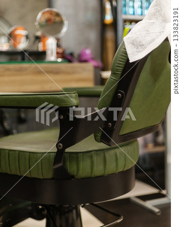 A vintage-style barber chair with green upholstery sits in a trendy barbershop. The clean interior features bright lighting and various hair products on display, creating a welcoming vibe A vintage-style barber chair with green upholstery sits in a trendy barbershop. The clean interior features bright lighting and various hair products on display, creating a welcoming vibe 133823191