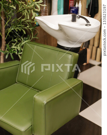A green salon chair is positioned next to a white washbasin in a barbershop. The space has a modern design with a mix of colors and textures, creating a welcoming atmosphere for clients A green salon chair is positioned next to a white washbasin in a barbershop. The space has a modern design with a mix of colors and textures, creating a welcoming atmosphere for clients 133823197