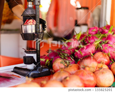 A fruit vendor uses a juicer to extract juice from bright pomegranates while surrounded by colorful dragon fruit. The cheerful setting enhances the freshness of the offerings A fruit vendor uses a juicer to extract juice from bright pomegranates while surrounded by colorful dragon fruit. The cheerful setting enhances the freshness of the offerings 133823201