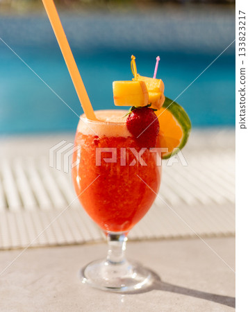 A vibrant cocktail blends tropical flavors, served in a clear glass. The drink is decorated with fruit and sits near a sparkling blue pool under the warm sun A vibrant cocktail blends tropical flavors, served in a clear glass. The drink is decorated with fruit and sits near a sparkling blue pool under the warm sun 133823217