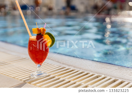 A refreshing tropical cocktail sits on the edge of a pool, adorned with fresh fruit and a colorful umbrella. Sunlight reflects on the water, creating a vibrant atmosphere A refreshing tropical cocktail sits on the edge of a pool, adorned with fresh fruit and a colorful umbrella. Sunlight reflects on the water, creating a vibrant atmosphere 133823225