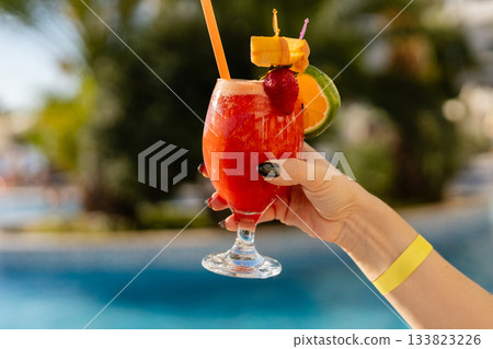 A hand holds a vibrant tropical cocktail near a sparkling pool, showcasing bright colors and garnishes, perfect for a sunny day of relaxation and enjoyment. All inclusive concept. 133823226