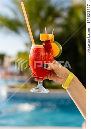 A hand holds a vibrant tropical drink adorned with fruit, enjoyed beside a sparkling blue pool under clear skies. The scene radiates summer vibes and relaxation. All inclusive concept. A hand holds a vibrant tropical drink adorned with fruit, enjoyed beside a sparkling blue pool under clear skies. The scene radiates summer vibes and relaxation. All inclusive concept. 133823227