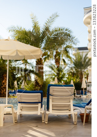 Two white lounge chairs with blue cushions sit beside a sparkling pool surrounded by tall palm trees and clear blue skies. A large umbrella provides shade, creating a serene oasis 133823228