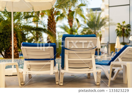 Comfortable white lounge chairs with blue cushions sit near a sparkling pool, surrounded by lush palm trees under a bright sun. Guests can enjoy a relaxing day outdoors. All inclusive. Comfortable white lounge chairs with blue cushions sit near a sparkling pool, surrounded by lush palm trees under a bright sun. Guests can enjoy a relaxing day outdoors. All inclusive. 133823229