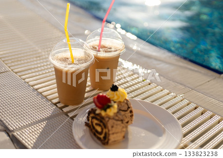 Two iced drinks with colorful straws sit beside a slice of cake on a plate. The setting is bright and sunny, with a pool in the background, creating a relaxing atmosphere for enjoyment 133823238