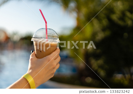 A hand holds a clear cup of iced coffee with a pink straw, set against a sunny backdrop of palm trees and a sparkling pool. Perfect for a warm day A hand holds a clear cup of iced coffee with a pink straw, set against a sunny backdrop of palm trees and a sparkling pool. Perfect for a warm day 133823242