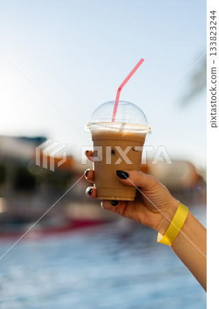 A hand holds a clear cup of iced coffee with a pink straw, set against the backdrop of a bright pool area. The scene captures a relaxing summer moment. All inclusive. 133823244