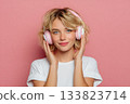 Young attractive blonde woman in white headphones and a white T-shirt listening to music on a pink background 133823714