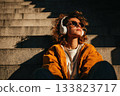 A young woman with curly hair wearing big white headphones and sunglasses sits on the steps on a sunny day 133823717