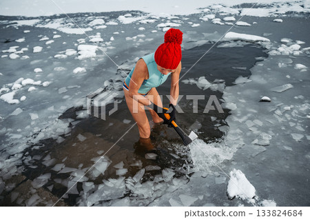 Senior woman breaking ice for winter swimming on frozen lake. 133824684