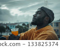 A Black man in athletic wear takes a moment to rest with a cup of tea in hand, seated in an urban outdoor space under an overcast sky. A scene blending wellness, style, and modern city life. 133825374