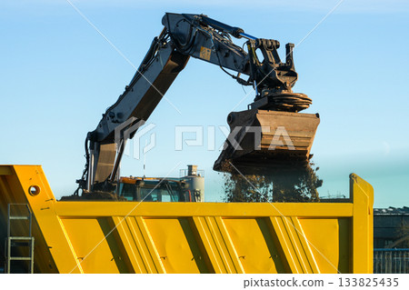 Excavator unloading soil into large yellow dump truck bed at construction site on clear sunny day Excavator unloading soil into large yellow dump truck bed at construction site on clear sunny day 133825435