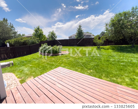 A clear summer day in a suburban backyard with a red wooden deck, lush grass, and a landscaped garden bed. Tall trees and a privacy fence create a peaceful and enclosed environment. A clear summer day in a suburban backyard with a red wooden deck, lush grass, and a landscaped garden bed. Tall trees and a privacy fence create a peaceful and enclosed environment. 133825605