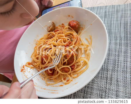 Close-up of a white bowl filled with spaghetti and meatballs in tomato sauce. A person wearing pink is holding the bowl on a woven placemat surface with a fork resting inside. Natural daylight creates Close-up of a white bowl filled with spaghetti and meatballs in tomato sauce. A person wearing pink is holding the bowl on a woven placemat surface with a fork resting inside. Natural daylight creates 133825615