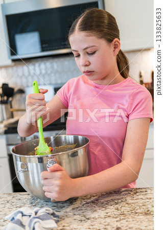 A girl in a pink t-shirt stirs a bowl of cookie dough using a green spatula. She is standing at a kitchen counter with granite surfaces and white cabinetry. A girl in a pink t-shirt stirs a bowl of cookie dough using a green spatula. She is standing at a kitchen counter with granite surfaces and white cabinetry. 133825633