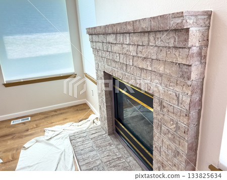 Side perspective showing a fully remodeled fireplace with brick pattern and gold-trimmed black insert. The environment includes window light and cloth on the floor. Side perspective showing a fully remodeled fireplace with brick pattern and gold-trimmed black insert. The environment includes window light and cloth on the floor. 133825634