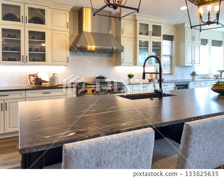 Wide view of a contemporary kitchen featuring a large island, pendant lights, and stainless steel appliances. The scene highlights open design, bright lighting, and elegant home details. Wide view of a contemporary kitchen featuring a large island, pendant lights, and stainless steel appliances. The scene highlights open design, bright lighting, and elegant home details. 133825635