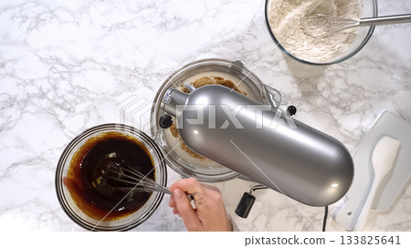 A hand is seen pouring dark vanilla extract into a glass bowl of cookie dough mixture on a marble surface. A whisk and a bowl of flour are nearby, emphasizing the baking preparation stage. Bright A hand is seen pouring dark vanilla extract into a glass bowl of cookie dough mixture on a marble surface. A whisk and a bowl of flour are nearby, emphasizing the baking preparation stage. Bright 133825641